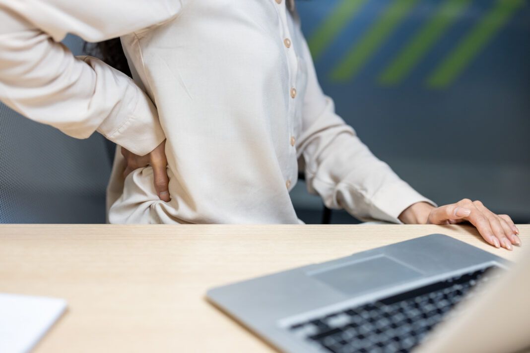Office worker experiences back pain while sitting at desk with laptop and presses on lower back for relief