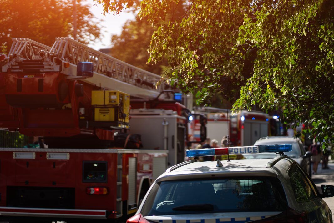 Firefighters and police respond to emergency, fire trucks with ladders and flashing blue lights. Firemen in gear prepare equipment in residential area